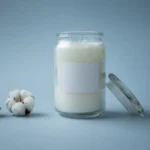 A minimalist shot of a glass jar candle beside cotton balls on a soft blue backdrop.